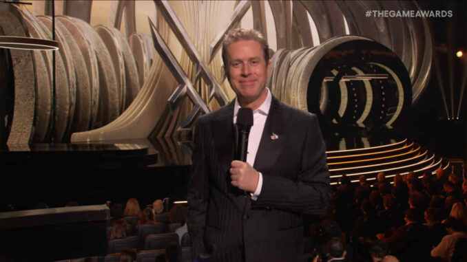 Geoff Keighley stojący na tle oprawy wydarzenia oraz przed osobami nominowanymi na miejscach siedzących.