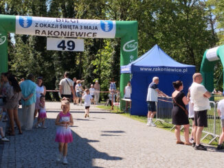 Uczestnicy majówki na tle parku, dmuchana brama START?META z napisem “BIEGI Z OKAZJI ŚWIĘTA 3 MAJA RAKONIEWICE 49”