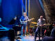 Zespół jazzowy występujący na żywo w zabytkowym, piwnicznym klubie z kamiennymi ścianami i łukowym sklepieniem. Scena jest nastrojowo oświetlona niebieskimi reflektorami. Od lewej do prawej widać: pianistę gitarzystę basowego, saksofonistę i perkusistę.