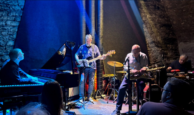 Zespół jazzowy występujący na żywo w zabytkowym, piwnicznym klubie z kamiennymi ścianami i łukowym sklepieniem. Scena jest nastrojowo oświetlona niebieskimi reflektorami. Od lewej do prawej widać: pianistę gitarzystę basowego, saksofonistę i perkusistę.