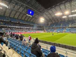 Zdjęcie zrobione z trybun stadionu piłkarskiego Lecha Poznań w nocy. Piłkarze rozgrzewają się na oświetlonej murawie.