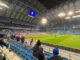 Zdjęcie zrobione z trybun stadionu piłkarskiego Lecha Poznań w nocy. Piłkarze rozgrzewają się na oświetlonej murawie.